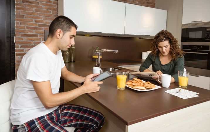 Couple sitting at kitchen island with newspaper, tablet, and pastries