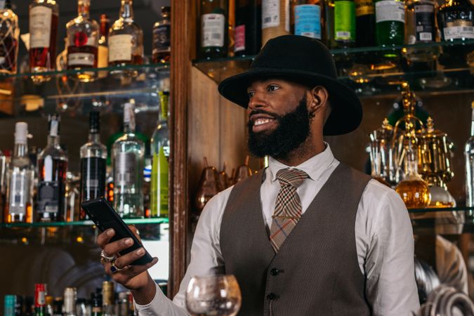 Stylish Black bartender in hat using smartphone