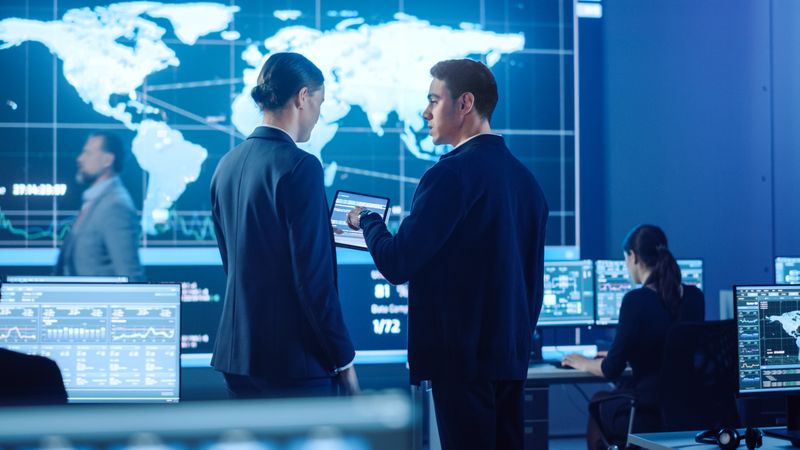 Professionals Review Data on Tablet in Futuristic Control Room. Large World Map Display Illuminates Background. Team Monitors Global Information on Multiple Screens in Blue-lit Environment.