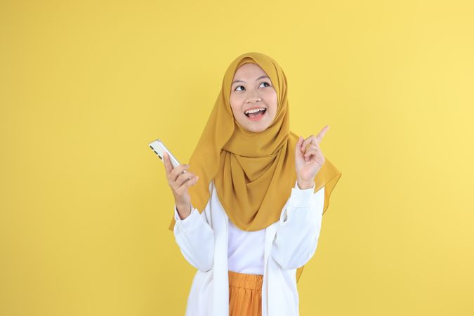 Look There, Excited Muslim Woman in Hijab Pointing Upwards and Holding Mobile Phone  at Copy Space