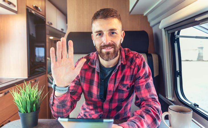 Man in red checkered shirt waving while sitting in van with digital tablet and coffee