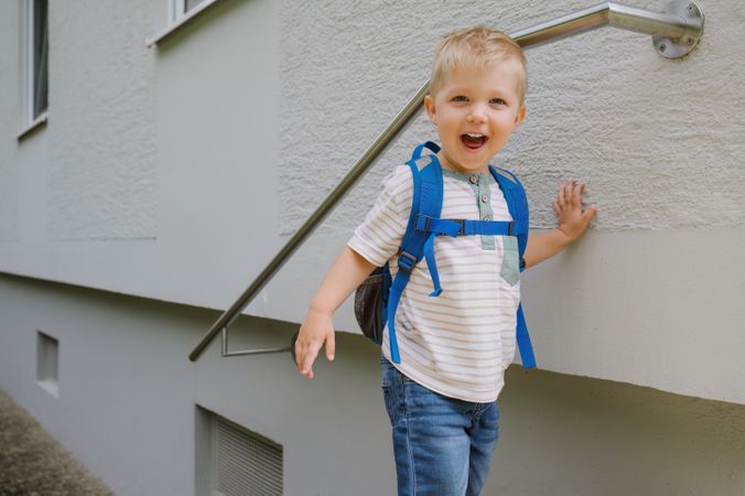Happy cute kid boy with backpack standing near gray wall building in daylight