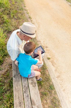 Grandchild and grandfather using a tablet outdoors on park bench, vertical