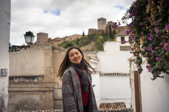 Asian female traveler in warm wear smiling among old buildings near big castle