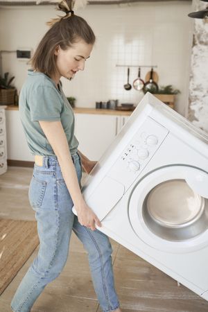 Joyful strong woman carrying washing machine in kitchen