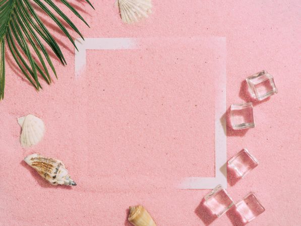 Pink sand with paper square outline and ice cubes, palm leaf and sea shells