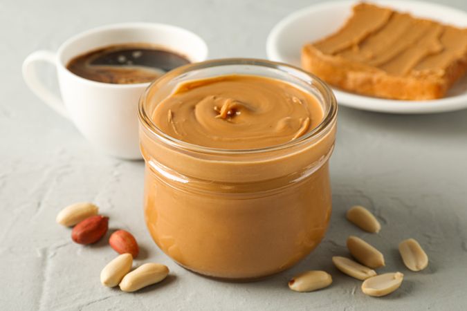 Glass jar with peanut butter, peanut, cup of coffee and peanut butter sandwich on grey background, closeup and space for text