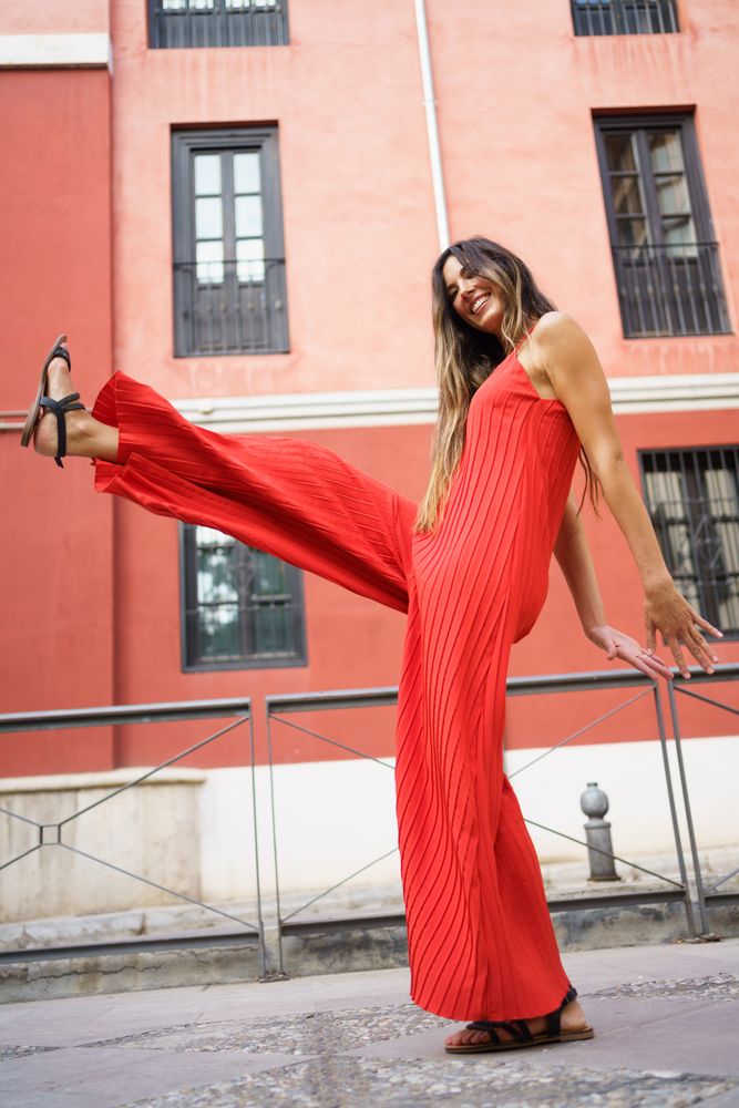 A woman in a vibrant red polka dot jumpsuit cheerfully poses outdoors in a bright setting