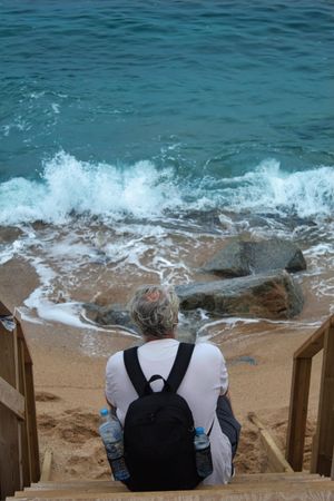Back view of middle aged man in light shirt sitting on wooden steps by the sea