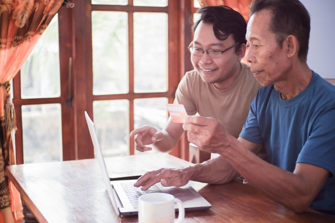 Happy adult son and older father pay online using laptop and credit card