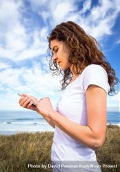 Woman in t-shirt checking phone with ocean waves in background, vertical 0yWZa4
