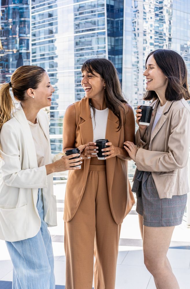 Three businesswomen are standing outside in front of a building, enjoying their office break, smiling and holding cups of coffee.