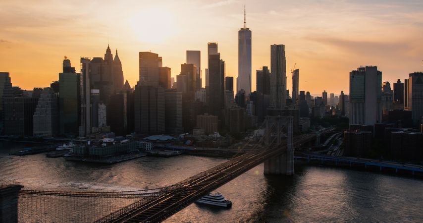 Iconic New York City Landscape Over East River with Skyscrapers, Brooklyn Bridge, Cars and Ferry Boats. Cinematic Evening Urban Skyline with Sunset and Lightly Clouded Sky