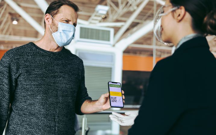 Male traveler in face mask showing his vaccine passport on smartphone to ground attendant