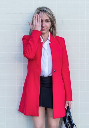 Portrait of a blonde woman wearing red jacket and skirt against a tiled wall covering her eye