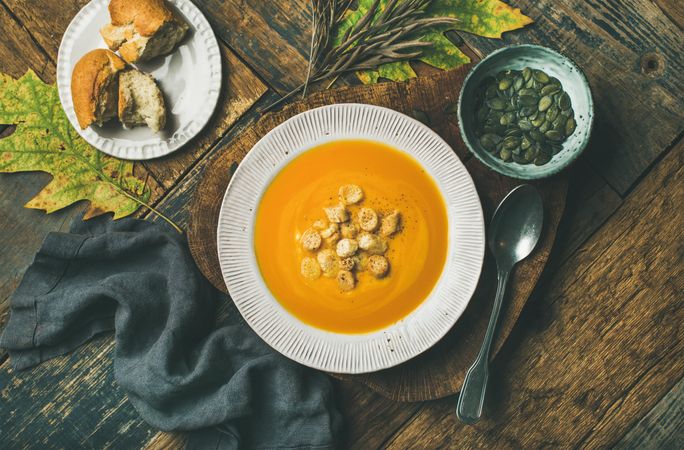Pumpkin soup with croutons, bread, seeds, spoon on wooden table with leaves