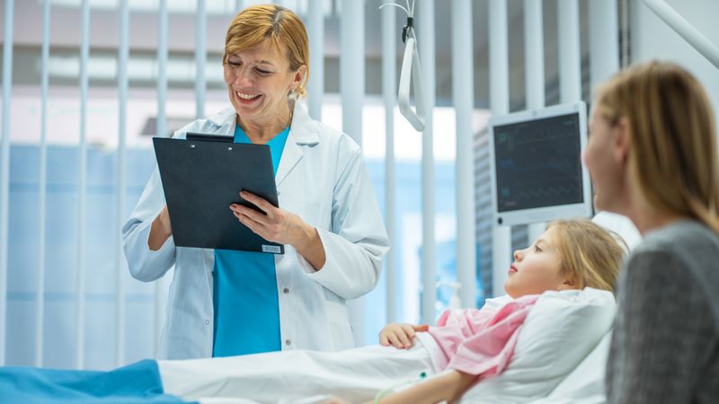 Recovering Little Girl Lies on a Bed In the Hospital, Friendly Doctor With Clipboard Asks Where it Hurts, Mother Sits Beside Bed