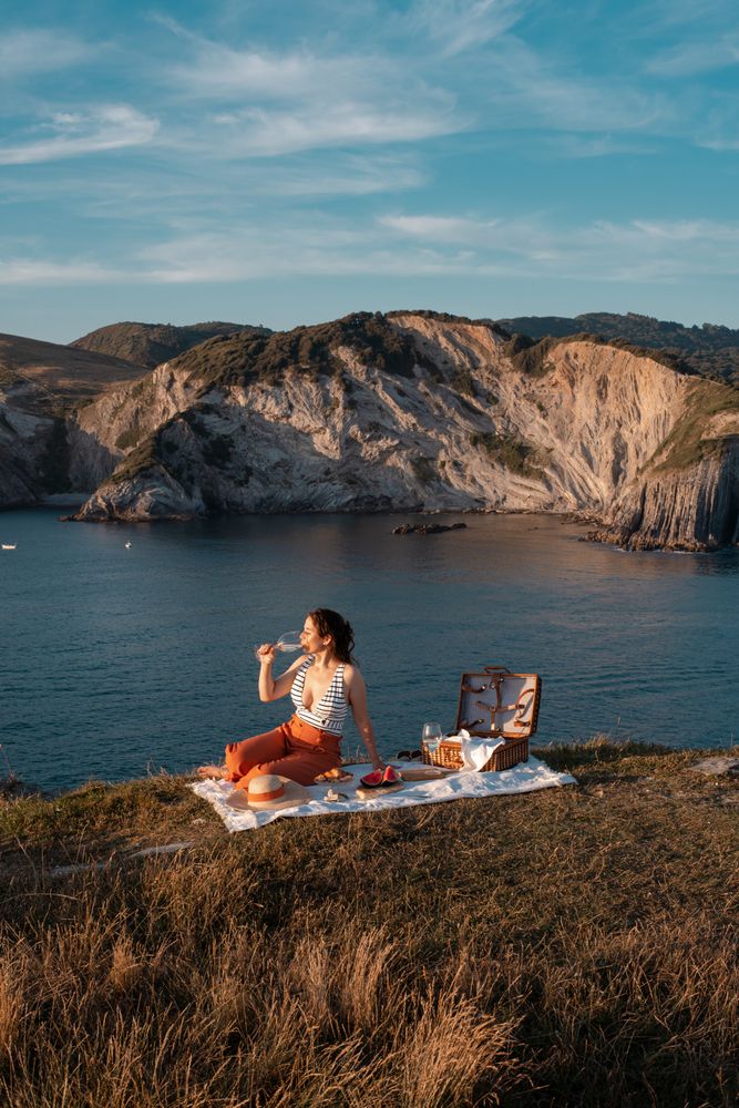 Woman with glass of drink on picnic mat looking at sea and mountains