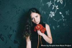Studio portrait of woman with blue eyes holding gerbera flower against dark floral wall 0yQ7O0