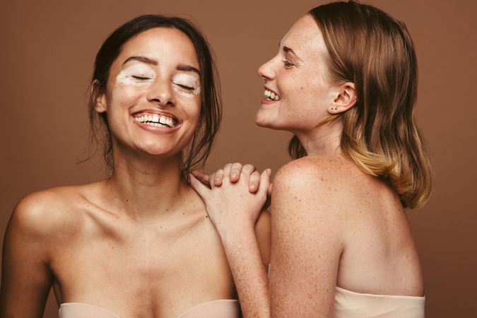 Two women with unique skin types smiling against brown background