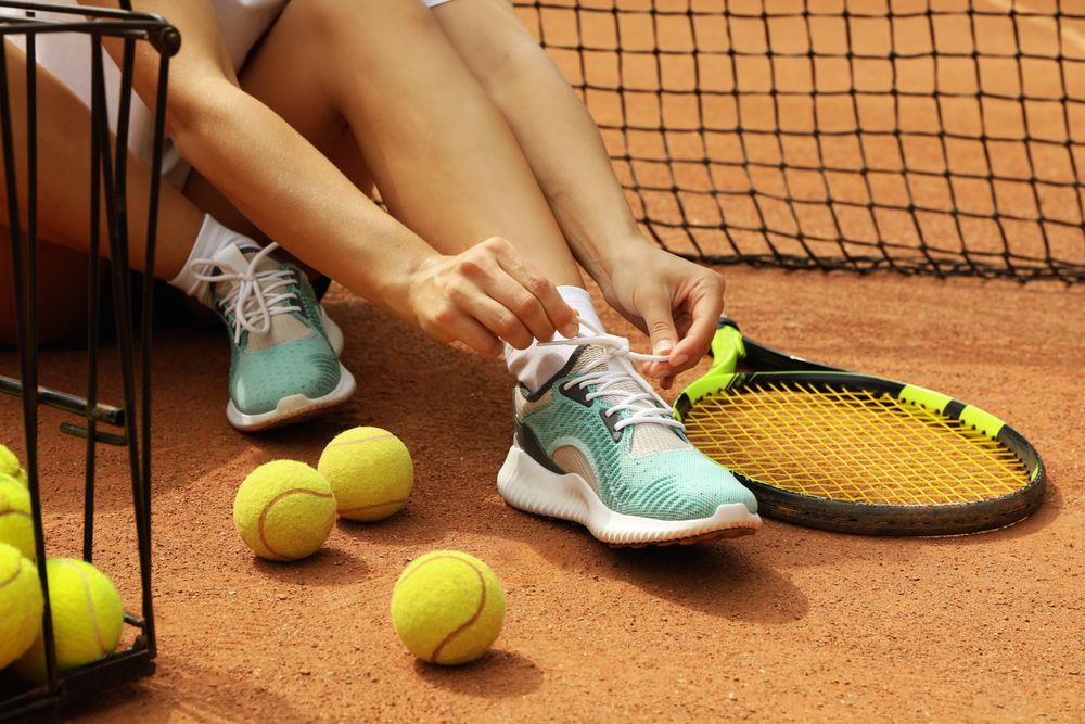 Woman tying shoelaces on clay court with racket and tennis balls