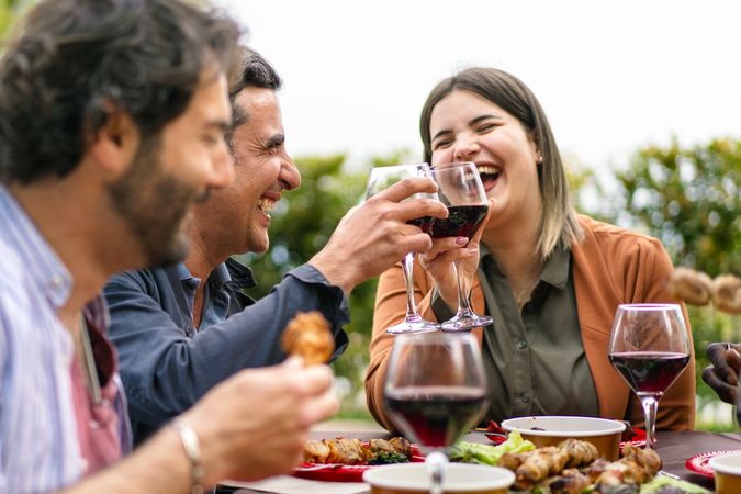 Joyful friends sharing a toast with red wine, laughing over a meal at an outdoor table surrounded by greenery