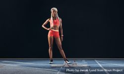 Female athlete standing beside a starting block on running track on a dark background 0JKPr5