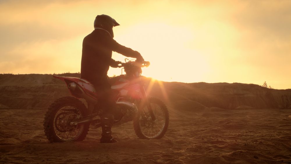 Professional FMX Motorcycle Rider Rests on His Bike and Overlooks Hard Sandy Off-Road Terrain.