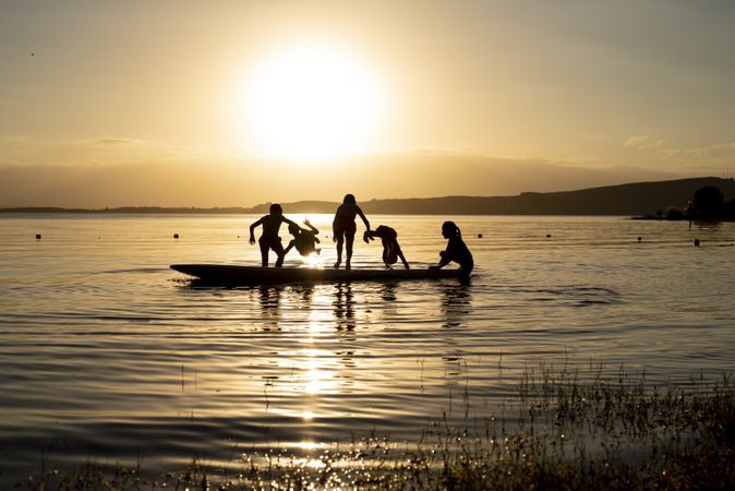 Silhouette of children playing and swimming around a surfboard on water during sunset