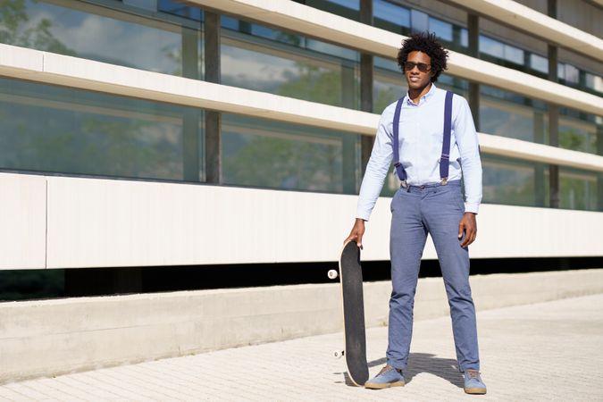 Cool man in sunglasses standing tall with skateboard outside building