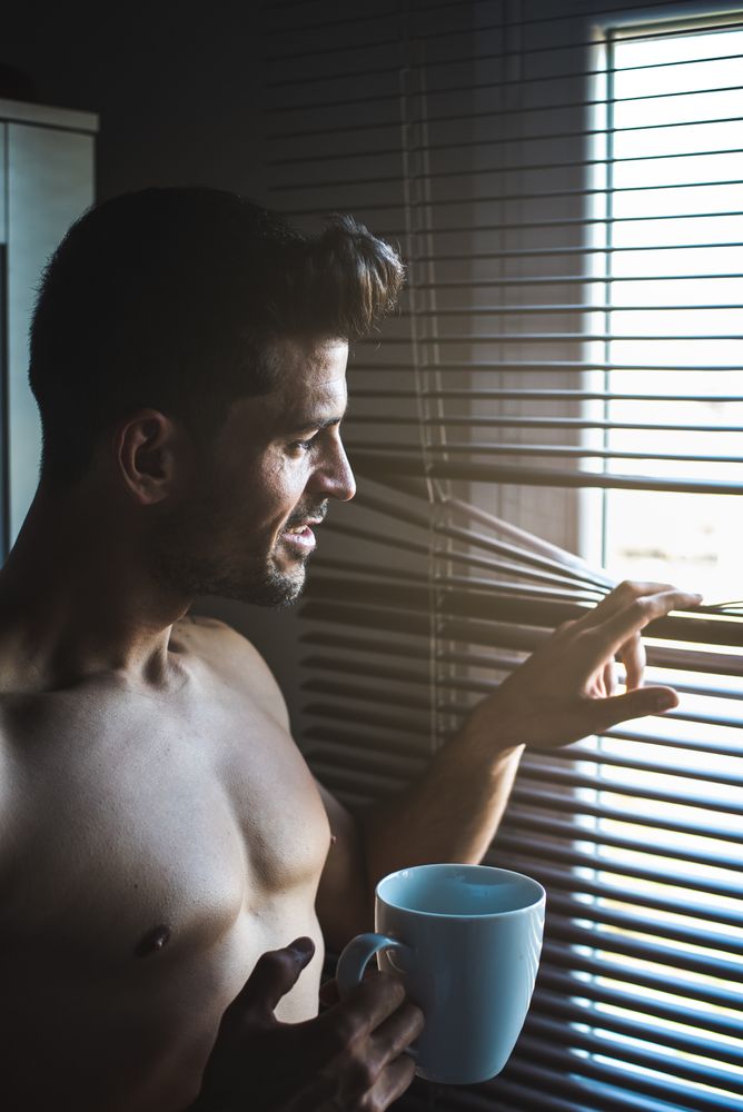 Man standing by a windows blind with a cup of coffee