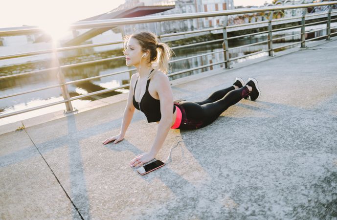 Woman with earphones and smartphone stretching on embankment