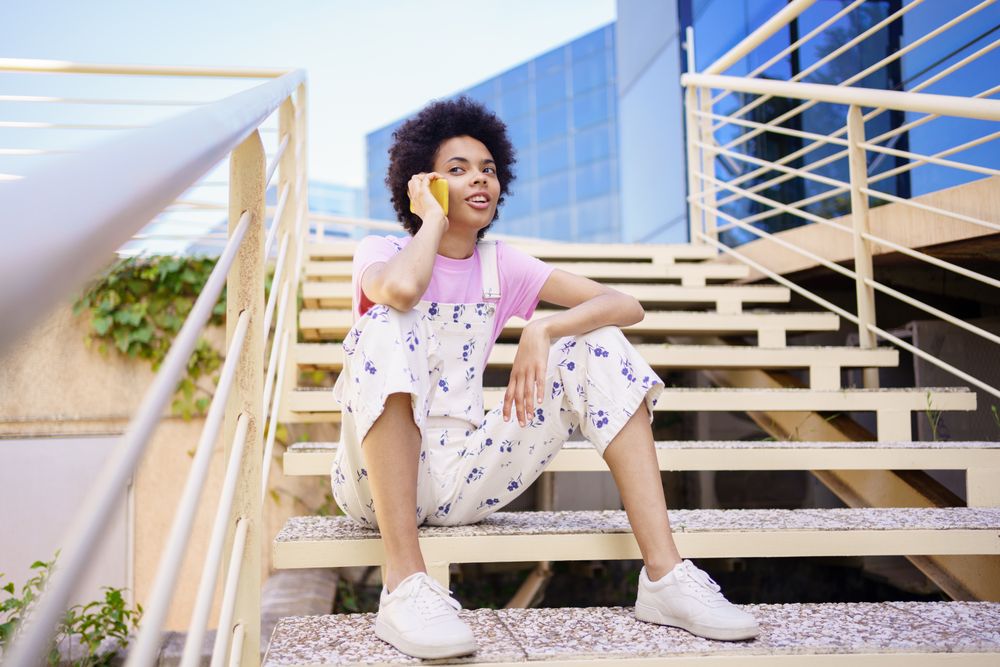 Bemused woman in floral overalls sitting on stairs taking phone call