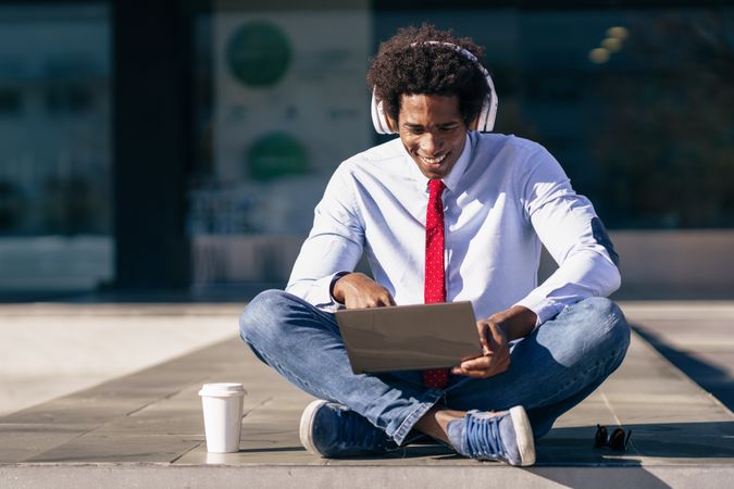 Smiling man sitting crosslegged outside with laptop, coffee and headphones
