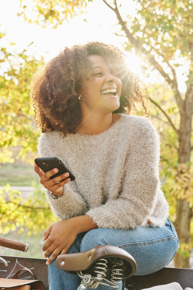 Laughing woman with curly hair using smartphone on bench in park