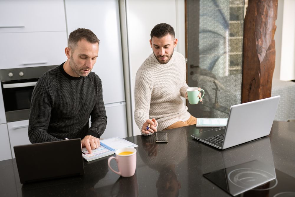 Two men with coffee working on laptops from their kitchen counter