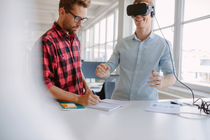 Young man writing notes with colleague wearing virtual reality goggles in office