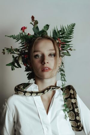 Young woman in light top with floral headdress and live snake on her shoulder against light background