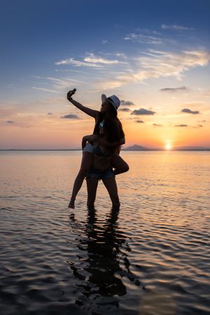 Woman carrying girlfriend on back and taking selfie in water at beach