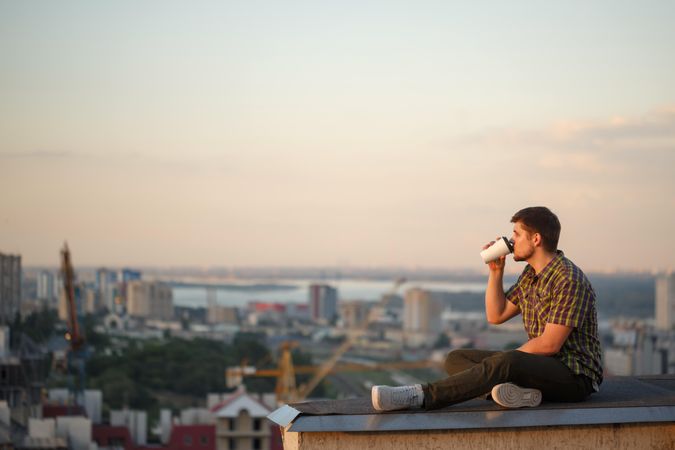 Side view of man perched on roof drinking coffee