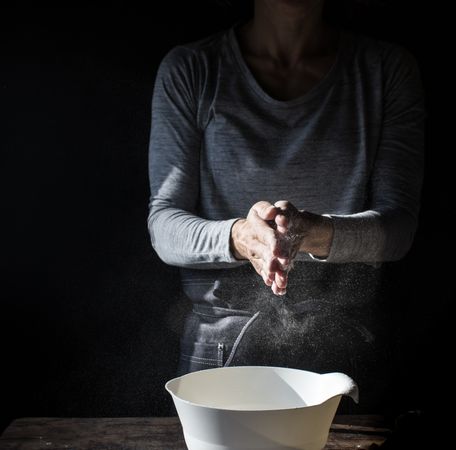 Woman clapping hands in flour near bowl and pack of eggs on table