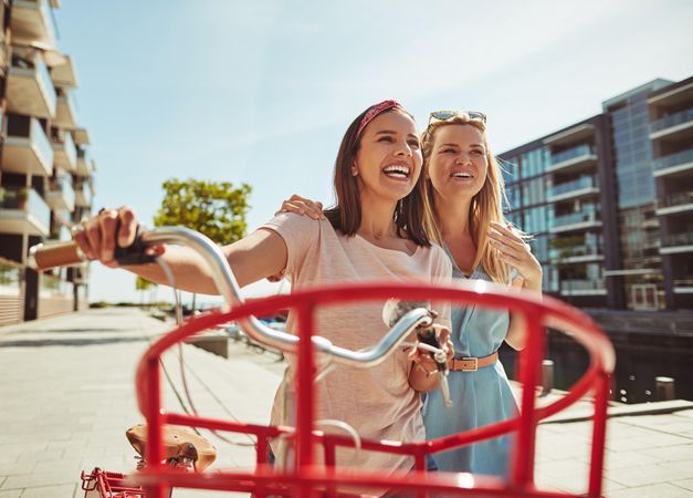 Two close female friends happily walking along a riverwalk with red bike on sunny day, close up