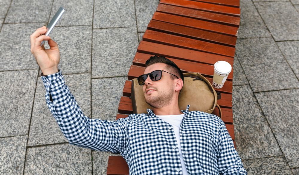Man lying on his back on a park bench making selfie with mobile