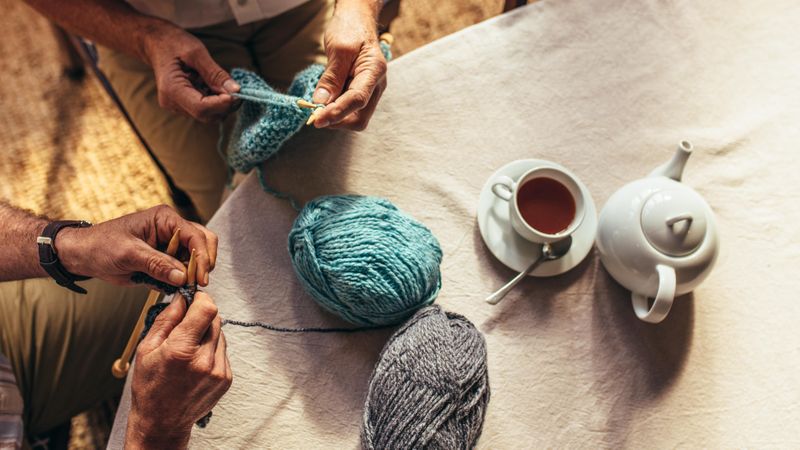 Top view of two men’s hands knitting with teapot and teacup on table