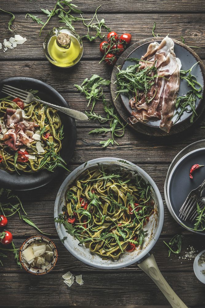 Italian food background with green cooked pasta in cooking pan with parma ham, arugula, tomatoes and cheese on rustic wooden kitchen table.