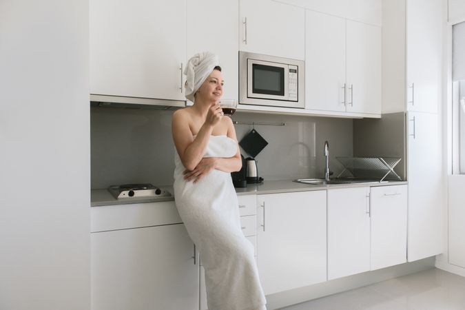Joyful adult woman enjoying day off while having hot drink after bath in apartment with simple interior design