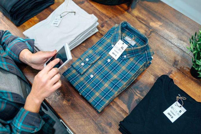 Unrecognizable female entrepreneur checking stock on phone next to apparel stacks on clothes store