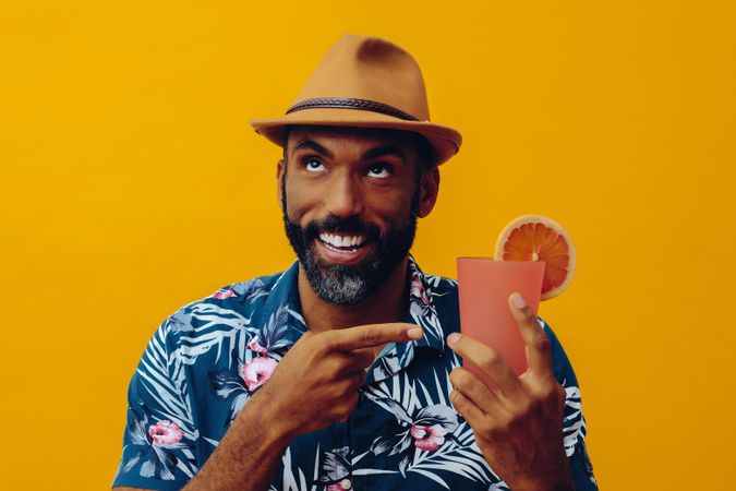 Happy male in hat and colorful shirt pointing to cocktail with yellow background
