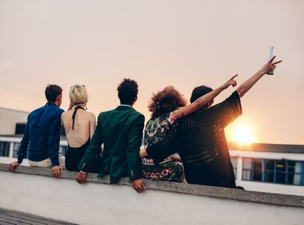 Young men and women enjoying drinks on rooftop at sunset with two pointing up
