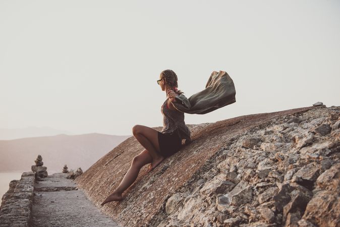 Woman sitting on castle wall overlooking sea, with wind blowing shirt